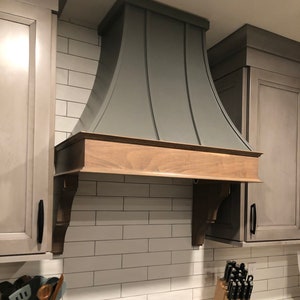 May include: A gray metal range hood with a wood trim, mounted above a black gas stove with a white countertop. The hood has a curved design and is installed in a kitchen with white subway tile backsplash.