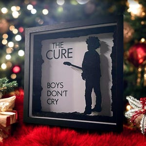 May include: A black framed shadow box with a white background featuring a silhouette of a person holding a guitar. The text "THE CURE" is above the silhouette and "BOYS DON'T CRY" is below the silhouette.