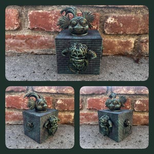 May include: A bronze-coloured decorative box featuring a whimsical creature on top and a grotesque face with ring handles on the front. The box has a brick-like texture, set against a brick wall background.