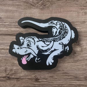 May include: A black and white alligator-shaped foam sticker with a light blue body and a pink tongue. The alligator is smiling and has detailed scales. The sticker is on a wooden surface.
