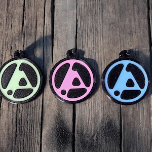 Op de afbeelding: Drie ronde zwarte sleutelhangers met een wit driehoeksontwerp. De driehoeken zijn gevuld met verschillende kleuren: groen, roze en blauw.