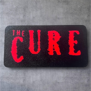 Könnte beinhalten: Ein rechteckiges schwarzes Schild mit den Worten "THE CURE" in leuchtend roter, verwitterter Schrift. Das Schild hat eine strukturierte, glitzernde Oberfläche. Der Text ist fett und in einer leicht gotischen Schriftart gehalten.