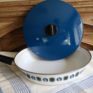 May include: White enamel frying pan with a blue lid. The pan has a black handle and a decorative floral pattern in green and blue around the rim. The lid is a solid, glossy blue. The pan is sitting on a striped cloth.