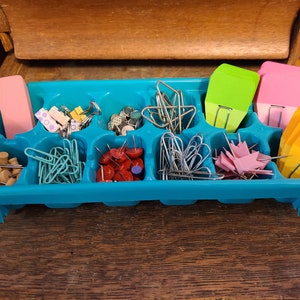 May include: A teal organizer filled with various office supplies. Includes a pink eraser, patterned push pins, paper clips, thumbtacks, and colorful sticky notes. The items are arranged in individual compartments.
