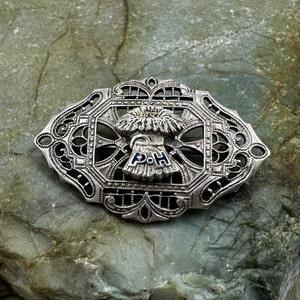 May include: Silver filigree brooch with a blue and white enamel design. The center features a stylized flower with the letters "P.O.H." in the center.