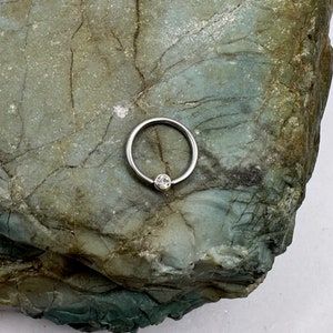 Puede incluir: Un anillo de plata con una piedra preciosa transparente, sobre un fondo texturizado verde y gris.