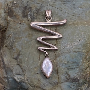 May include: A silver pendant with a wavy design and a white pearl drop. The pendant is on a gray rock background.