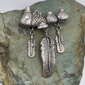 May include: Silver pendant featuring five charms: four small, detailed, silver pots and one larger, detailed, silver feather. The pots have a textured, geometric design. The feather has a detailed, realistic design.