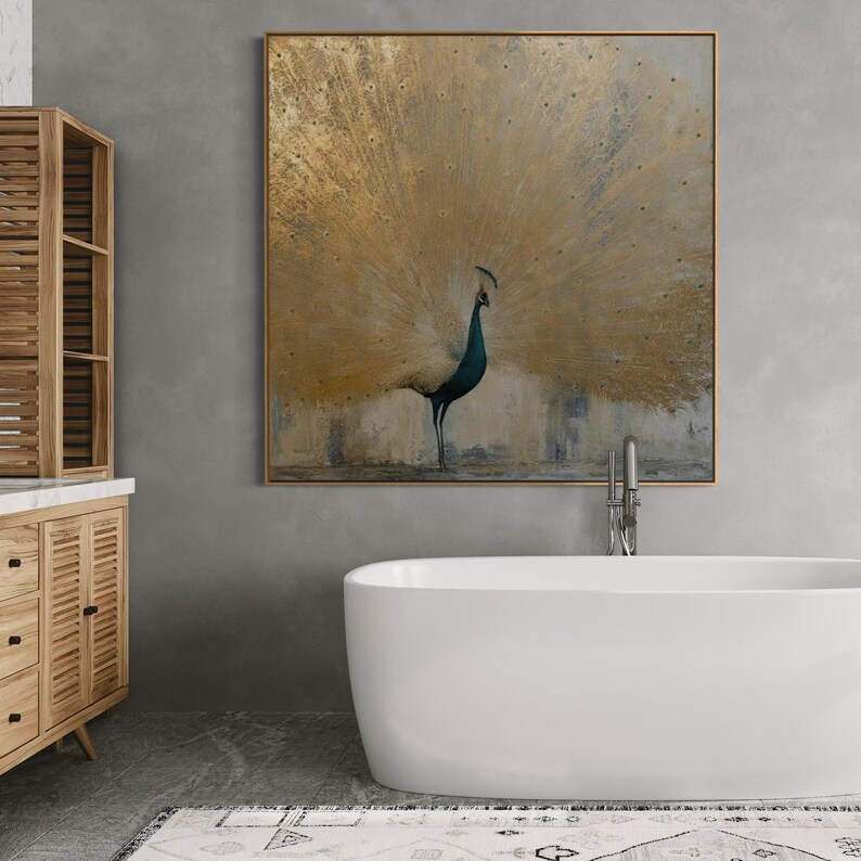 Op de afbeelding: Een vierkant wandkunstwerk met een pauw met een grote, uitgewaaierde staart in goud- en metallic bronskleuren. De vogel heeft een donkerblauwe nek en kop, geplaatst tegen een getextureerde, neutraal gekleurde achtergrond in een dunne houten lijst.