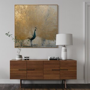 Op de afbeelding: Een vierkant wandkunstwerk met een pauw met een uitgewaaierde staart in goud- en wittinten. De vogel heeft een donkerblauwe nek en kop. Het kunstwerk is ingelijst in een dunne gouden rand en hangt boven een houten dressoir met een lamp.