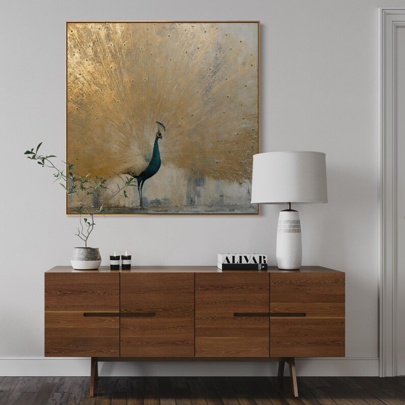 Op de afbeelding: Een vierkant wandkunstwerk met een pauw met een uitgewaaierde staart in goud- en wittinten. De vogel heeft een donkerblauwe nek en kop. Het kunstwerk is ingelijst in een dunne gouden rand en hangt boven een houten dressoir met een lamp.
