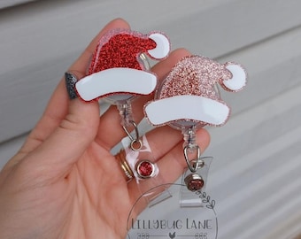 Può includere: Due porta badge a forma di cappello di Babbo Natale. Uno è rosso glitterato, l'altro è oro rosa glitterato, entrambi con finiture bianche. I porta badge hanno una base in plastica trasparente con una gemma rossa decorativa. Il logo Lillybug Lane è visibile.