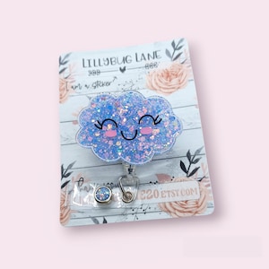 K&ouml;nnte beinhalten: Eine blaue Glitzerwolke mit einem l&auml;chelnden Gesicht auf wei&szlig;em Hintergrund. Die Wolke ist an einem einziehbaren Ausweishalter mit einem silbernen Clip befestigt. Der Text "Lillybug Lane" ist auf der Karte sichtbar.