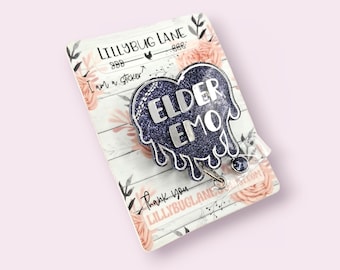 Puede incluir: Un portatarjetas en forma de corazón azul marino brillante con las palabras "ELDER EMO" en letras plateadas. El portatarjetas está sobre una tarjeta con estampado floral con el texto "LILLYBUG LANE" y "I am a sticker".