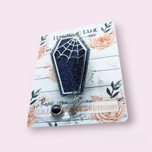 May include: A coffin-shaped badge reel with a glittery navy blue finish and a white spiderweb design. The badge reel is attached to a clear plastic holder with a metal clip. The background features a floral design and the text "LILLIBUG LANE".