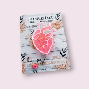 May include: Pink glitter anatomical heart shaped badge reel with a silver glitter outline. The reel is attached to a card with the text "Lillybug Lane" and "I am a sticker".