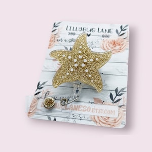 May include: A gold glitter starfish badge reel with white polka dots. The reel is attached to a decorative card with the text "LILLYBUG LANE" and "LILLYBUG LANE20.ETSY.COM". The background is a light pink color.