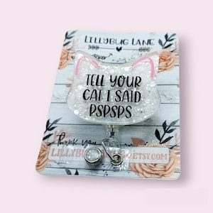 May include: A white glitter cat-shaped retractable badge holder with pink ears. The holder has the text "Tell your cat I said PSPSPS" printed on it. The holder is attached to a silver retractable badge reel.