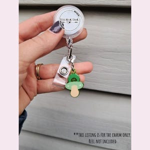 May include: A white retractable badge reel with a clear plastic strap and a charm. The charm is a green and tan pacifier shape with glitter accents. The reel has the text "Lillybug Lane" in a circular design.