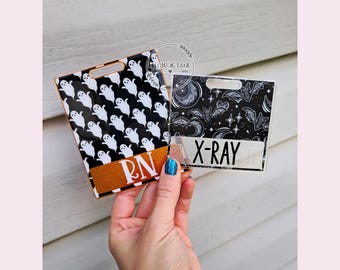 Pode incluir: Dois porta-crachás com tema de Halloween. Um apresenta fantasmas brancos em um fundo preto com "RN" em uma faixa dourada com glitter. O outro tem um design celestial com "X-RAY" em uma faixa branca. Ambos têm uma sobreposição de plástico transparente.