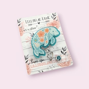 May include: A light blue manatee badge reel with a floral design, glitter accents, and a retractable cord. The badge reel is attached to a decorative card with the text "LILLYBUG LANE" and "Thank you".