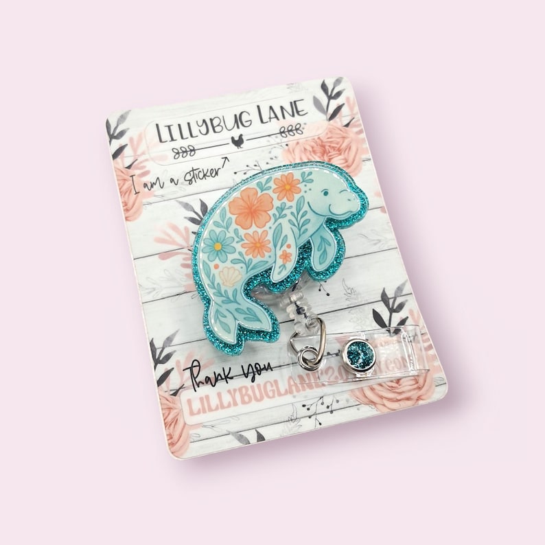 May include: A light blue manatee badge reel with a floral design, glitter accents, and a retractable cord. The badge reel is attached to a decorative card with the text "LILLYBUG LANE" and "Thank you".