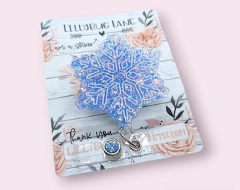 May include: A glittery blue snowflake badge reel with a silver clip. The badge reel is on a decorative card with the text "LILLYBUG LANE" and "Thank you". The snowflake design is detailed and sparkly.