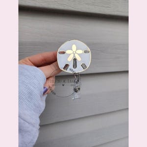 May include: A white and gold sand dollar charm with a flower design attached to a clear retractable badge holder.