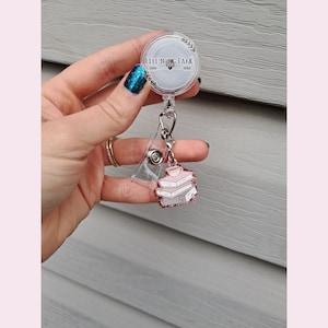 May include: A clear retractable badge reel with a stack of pink books and a teacup charm. The reel has the text "LILLYBUG LANE" and a floral design. The badge reel is attached to a clear plastic clip.
