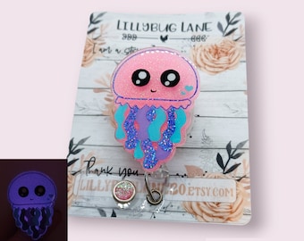 May include: Pink glitter resin jellyfish with blue and purple tentacles, a smiling face, and two small hearts. The jellyfish is attached to a clear badge holder with a silver retractable reel. The background is a white card with the text "Lillybug Lane" and "Thank you".