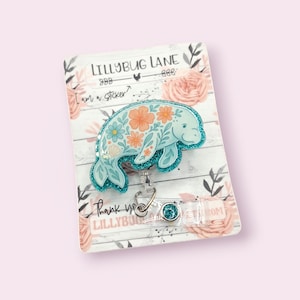May include: A light blue manatee badge reel with a floral design in orange and blue. The badge reel has a glittery border and the text "LILLYBUG LANE" and "I am a sticker?" is visible. The background is a floral design.