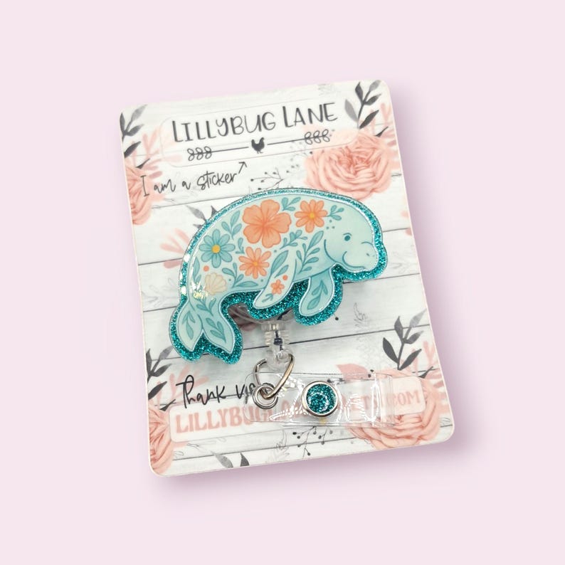 May include: A light blue manatee badge reel with a floral design in orange and blue. The badge reel has a glittery border and the text "LILLYBUG LANE" and "I am a sticker?" is visible. The background is a floral design.