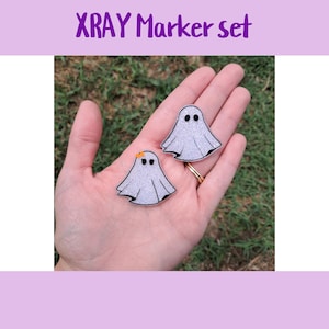 May include: Two glittery ghost-shaped XRAY marker set. One ghost has a small orange bow. The markers are held in a hand against a green background. The text "XRAY Marker set" is in purple.