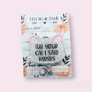 May include: A white glitter cat-shaped badge holder with pink ears and the text "TELL YOUR CAT I SAID PSPSPS".