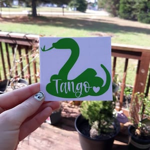 May include: A green vinyl sticker featuring a stylized snake design. The word "Tango" is written in a script font below the snake, accompanied by a small heart. The sticker is held against a background of greenery and a wooden deck.