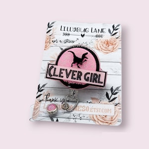 May include: Pink glitter badge reel with a black dinosaur silhouette and the text "Clever Girl". The reel is attached to a white card with the text "Lillybug Lane" and a floral design.