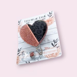 May include: A heart-shaped badge reel with a rose gold glitter finish and a black leopard print design. The badge reel is attached to a white card with floral accents and the text "LILLYBUG LANE". The website address etsy.com is also visible.