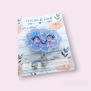 May include: A blue glitter cloud with a smiling face on a white card with the text "Lillybug Lane" and "I'm a sticker". The cloud is attached to a clear retractable badge holder.