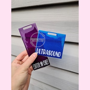 May include: Two rectangular acrylic tags with top slots. One is a deep purple tag with the white text EDITOR-IN-CHIEF. The other is a bright blue tag with the white text ULTRASOUND. Both tags feature a glossy finish and a circular logo.