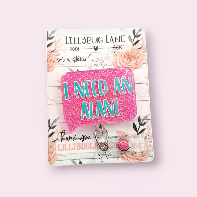May include: A pink glitter badge reel with the text "I NEED AN ALANI" in teal. The reel is attached to a decorative card with floral and wood grain elements. The card also features the text "LILLYBUG LANE" and "Thank you".