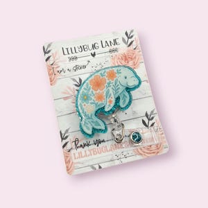 May include: A light blue manatee badge reel with orange and blue floral details. The badge reel has a glittery teal border and is attached to a clear plastic holder. The backing card has the text "LILLYBUG LANE" and "I am a sticker?"