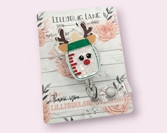 May include: A decorative badge reel with a baby bottle design and reindeer antlers. The bottle is white with red and green accents, and filled with glitter. The badge reel has a clear plastic strap and a metal clip.