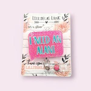 May include: A pink glitter badge reel with the text "I NEED AN ALANI" in teal. The badge reel has a speech bubble shape and a retractable cord with a metal clip. The backing card has floral and leaf designs.