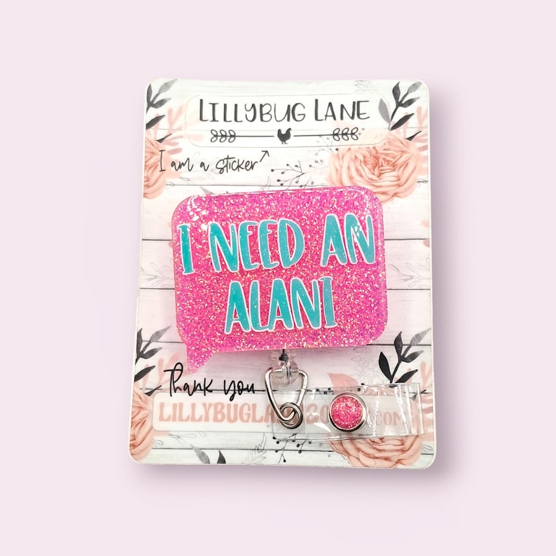 May include: A pink glitter badge reel with the text "I NEED AN ALANI" in teal. The badge reel has a speech bubble shape and a retractable cord with a metal clip. The backing card has floral and leaf designs.