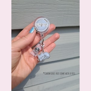 May include: A clear retractable badge reel with a silver-colored charm of a sleeping cat. The reel has the text "LITTLEBUG LANE" in a circular design. The charm is gray and white.