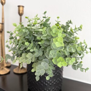 May include: A small artificial eucalyptus plant with light green, rounded leaves arranged on thin stems. The plant is housed in a matte black cylindrical pot featuring a textured, dotted surface pattern. Two tall, slender brass candle holders stand.