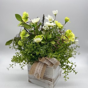 Puede incluir: Arreglo floral decorativo en una caja de madera blanqueada, con follaje verde, flores blancas y amarillas, y una cinta de arpillera y encaje. Tres huevos de Pascua moteados en tonos verdes y amarillos al frente.