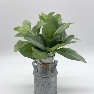 May include: A small, galvanized metal milk can vase filled with faux green leaves. The vase has two small handles and is wrapped with twine. The leaves are a mix of light and dark green, creating a natural look. The vase is set against a white background.