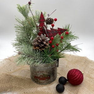 May include: A metal bucket with the text FRESH CUT CHRISTMAS TREES and a red truck graphic, filled with artificial pine branches, pinecones, red berries, and red plaid tree cutouts, accompanied by one large red and two small black ornaments.