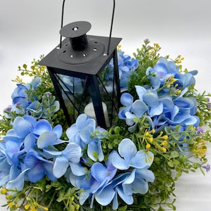 May include: A black metal lantern with a thin handle and a white candle inside, centered within a circular wreath of blue hydrangea flowers, green foliage, and small yellow accents. The lantern features a square frame and a vented top.
